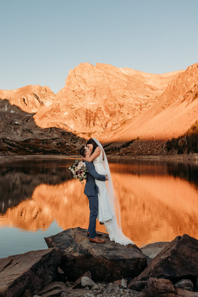 groom picks up bride under the butt in front of lake with sunrise reflected in it