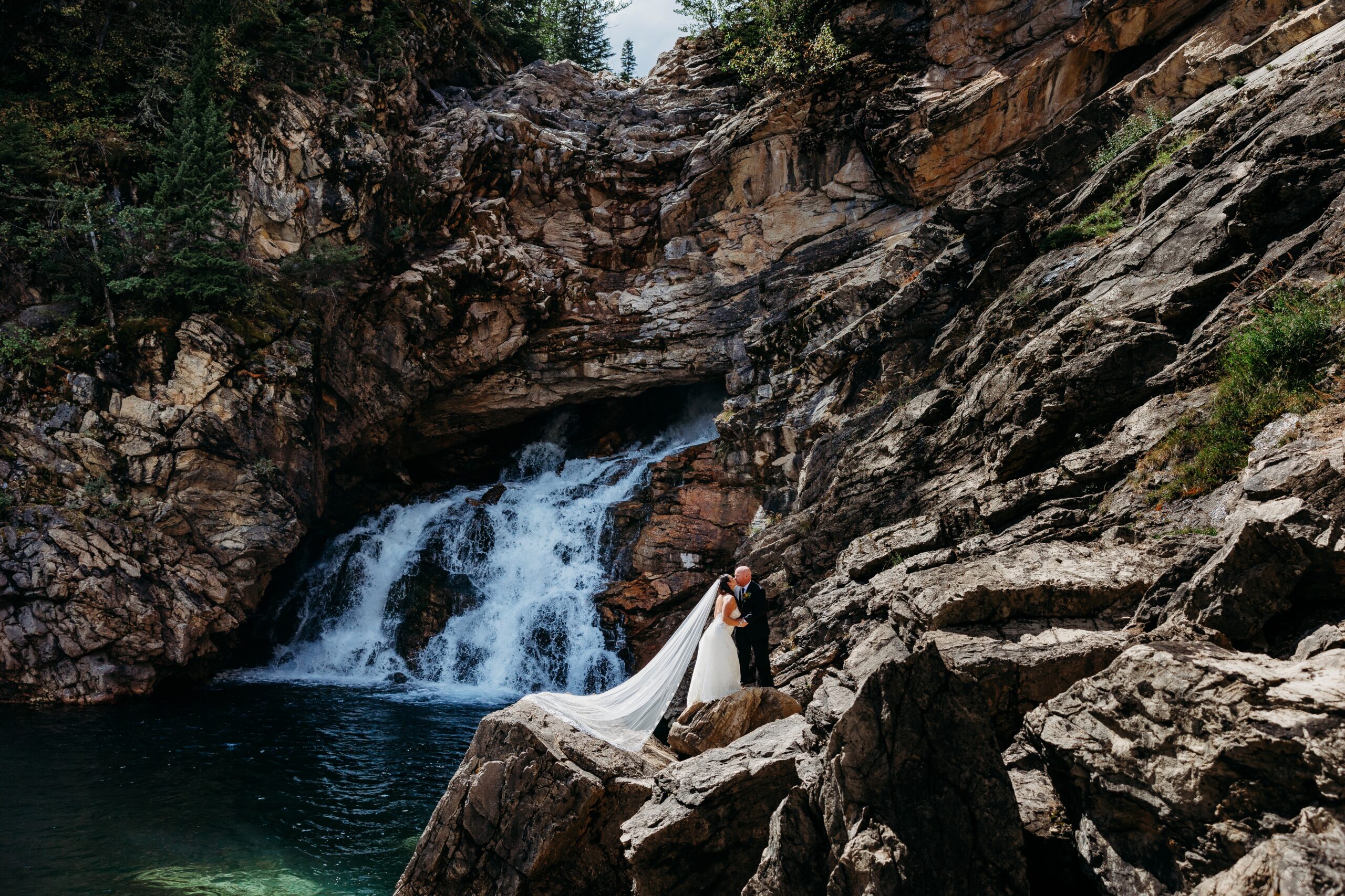 bride and groom stand on rocks in front of waterfall