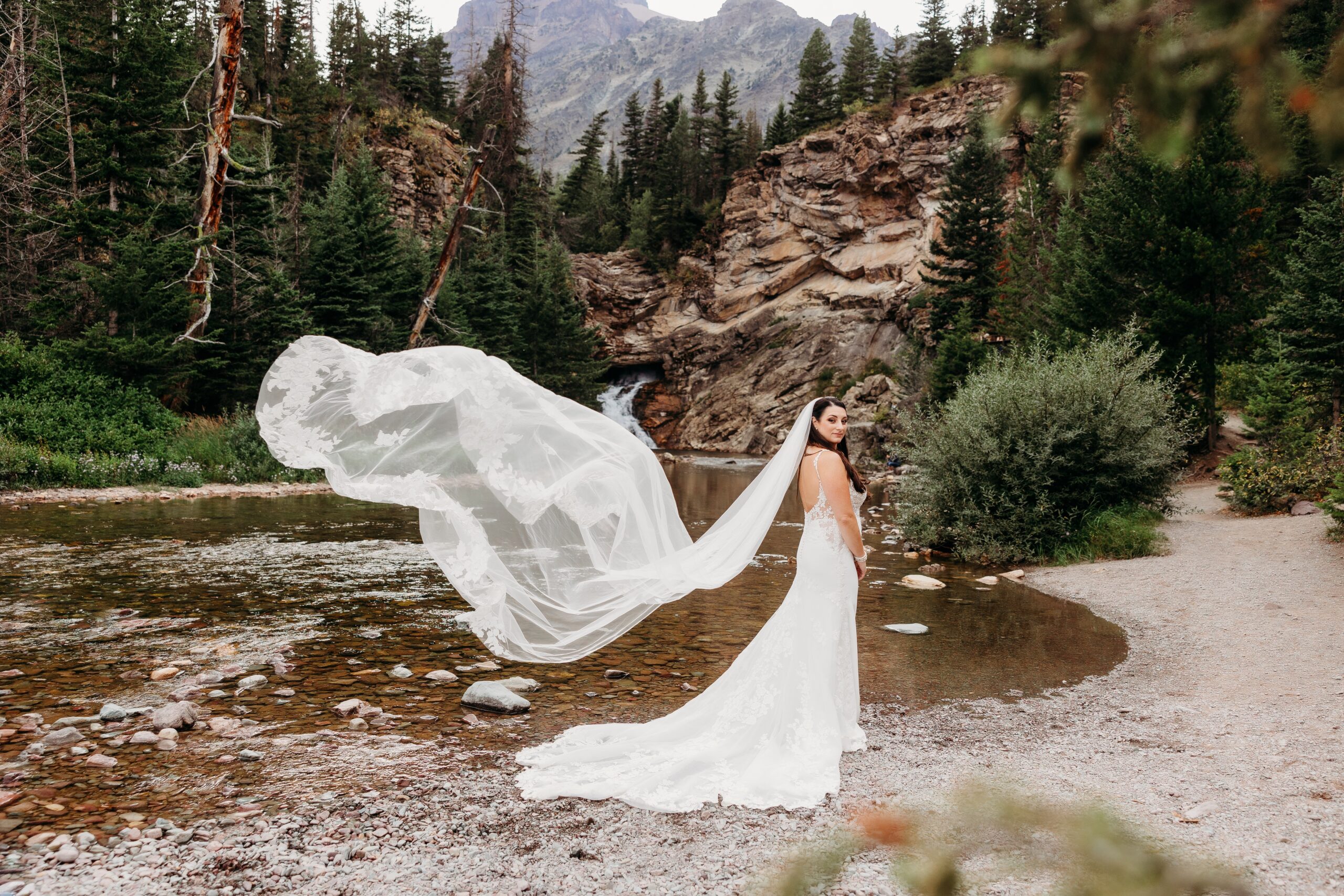 bride stands in front of river with veil flowing out behind her