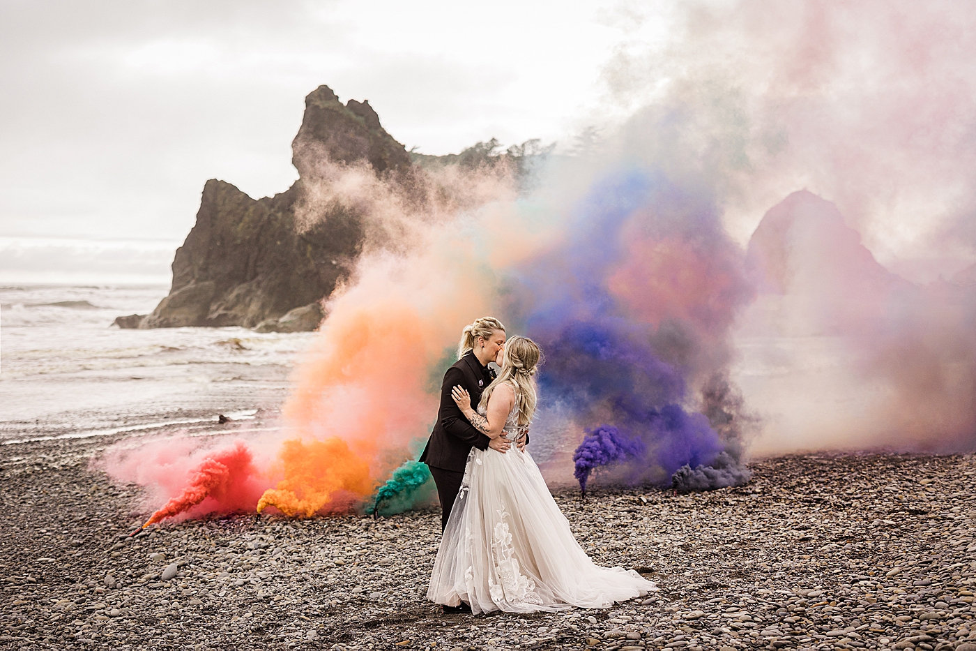 Bride's kissing after same-sex elopement at Ruby Beach with colorful smoke bombs in the background to celebrate pride month. Photo by Megan Montalvo Photography.
