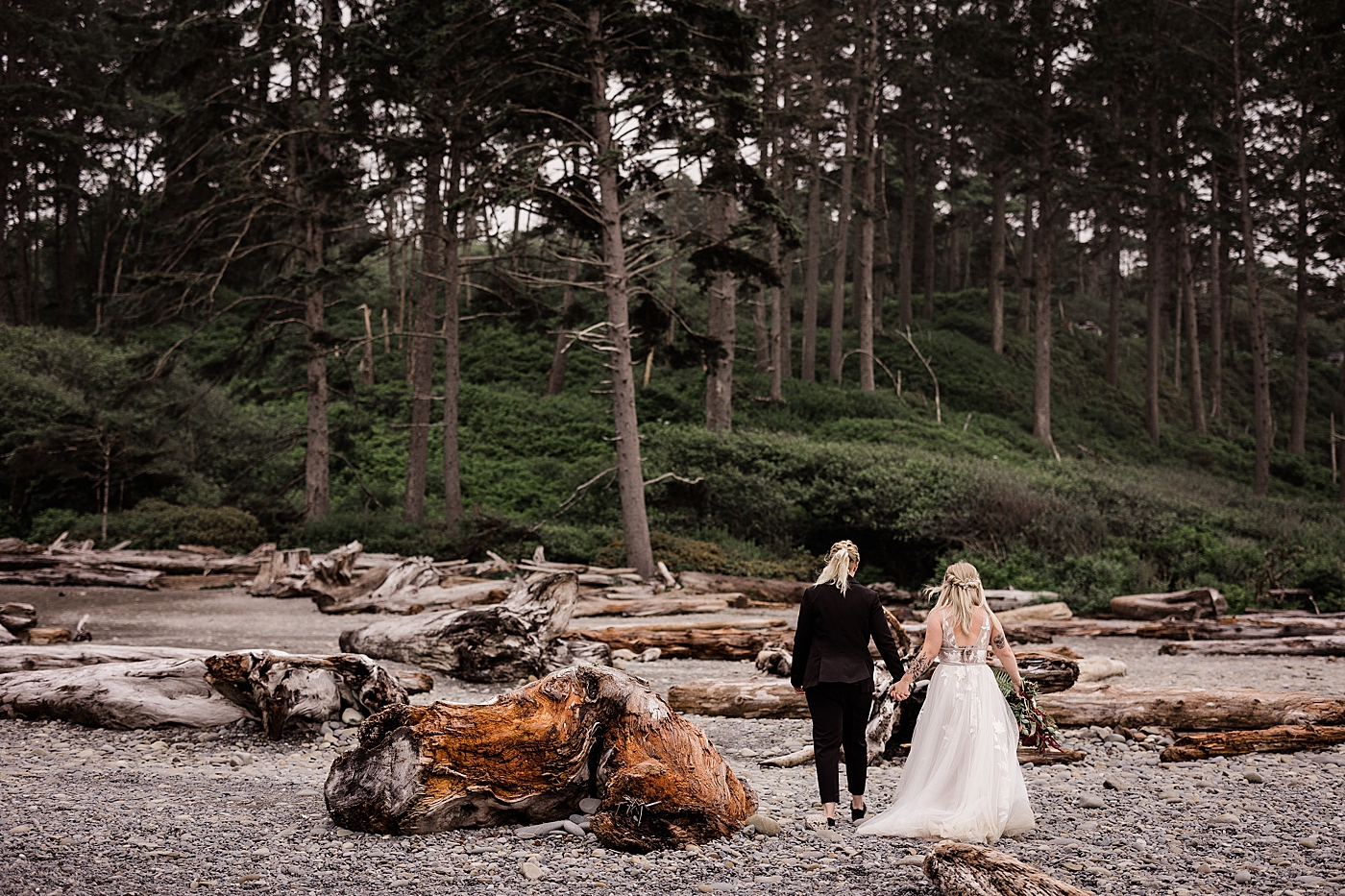 Couple holding hands walking along logs and trees at Ruby Beach. Photo by Megan Montalvo Photography.