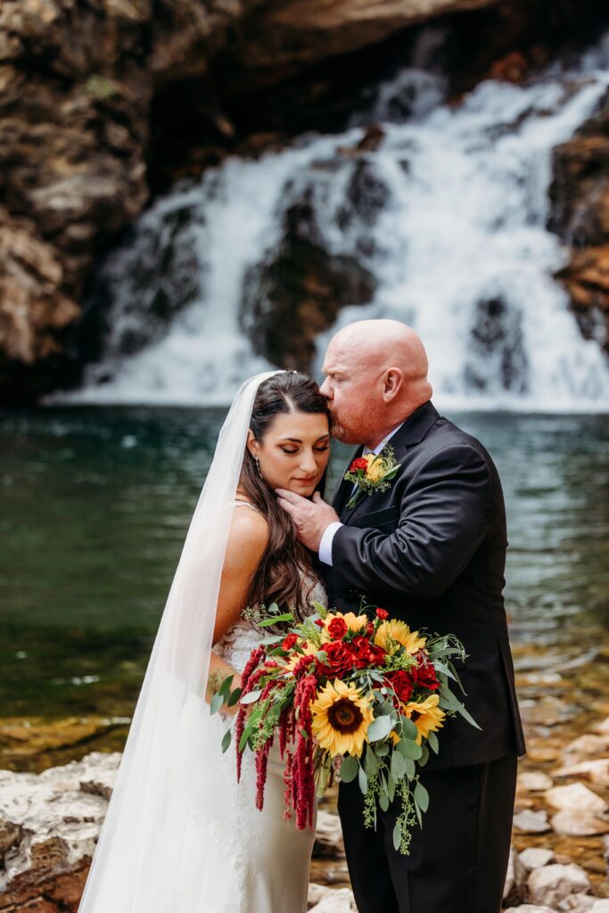 groom kisses bride on forehead in front of waterfall