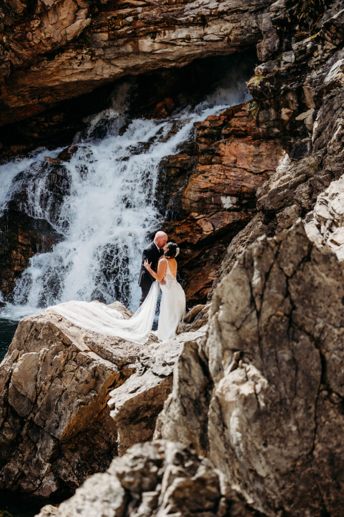 bride and groom stand on rocks in front of waterfall