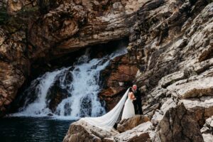 bride and groom stand on rock ledge looking out at waterfall