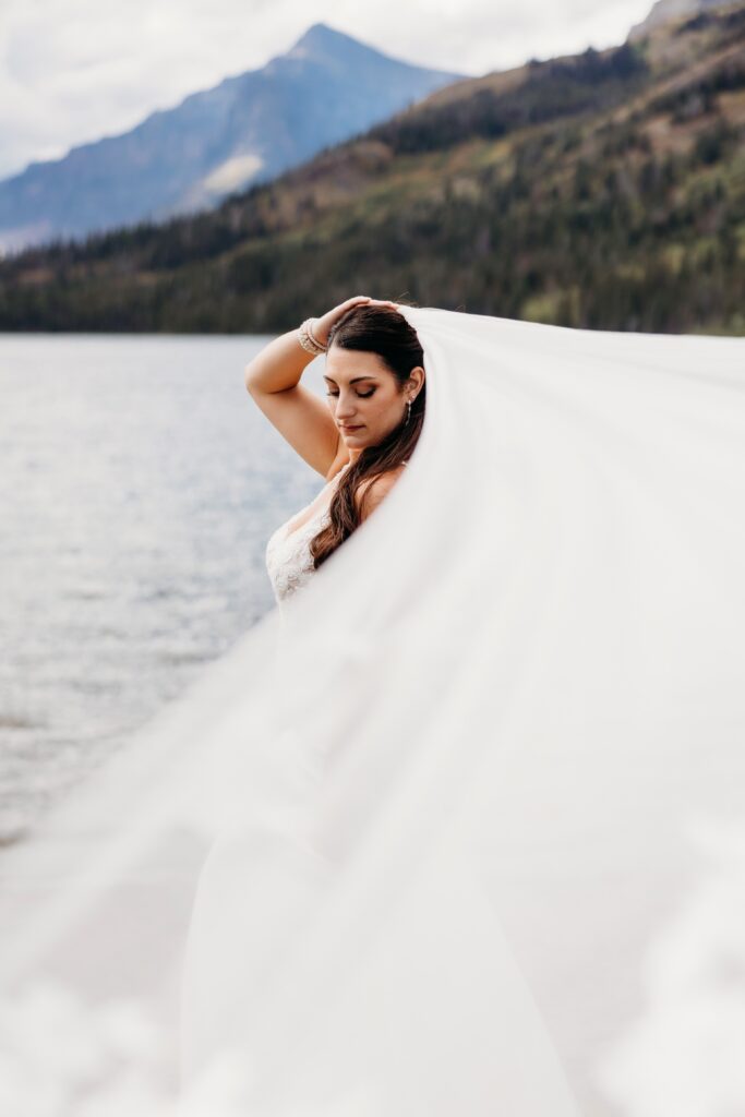 bride holds veil on head with lake behind her