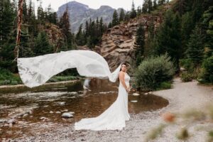bride stands in front of river with veil flowing out behind her