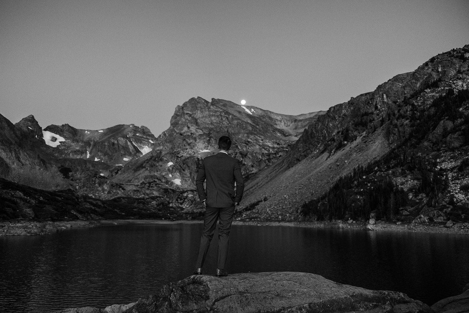 groom stands on edge of rock and looks out over lake