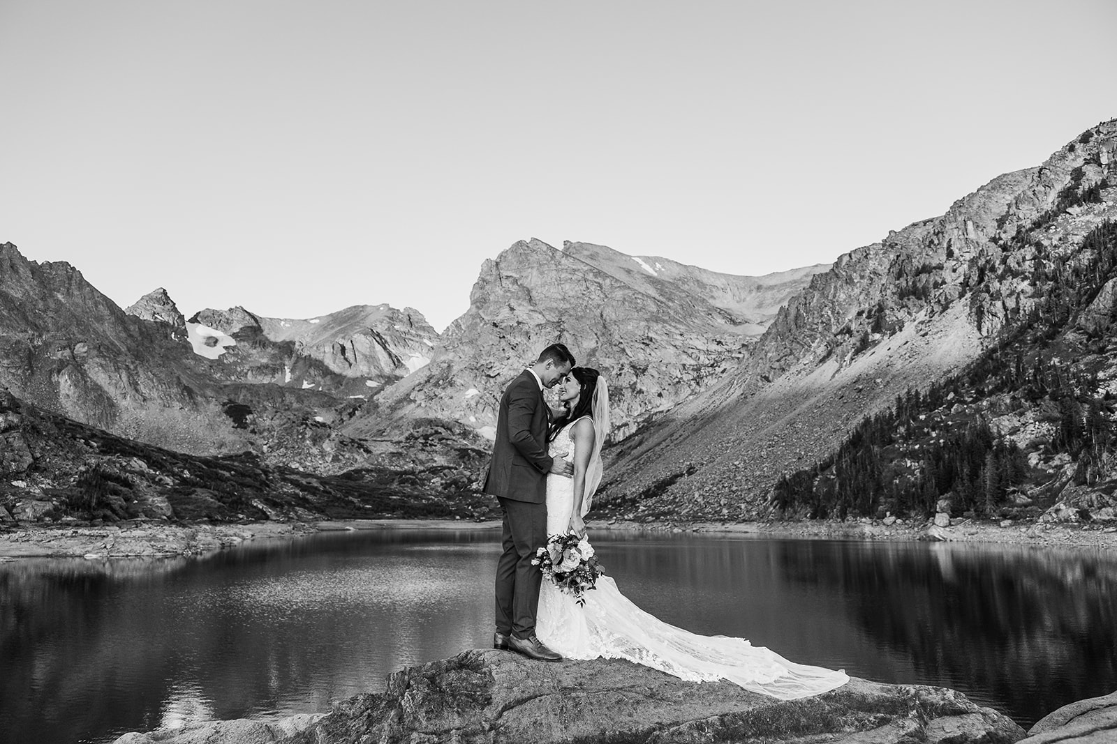 bride and groom embrace on a rock overlooking a lake
