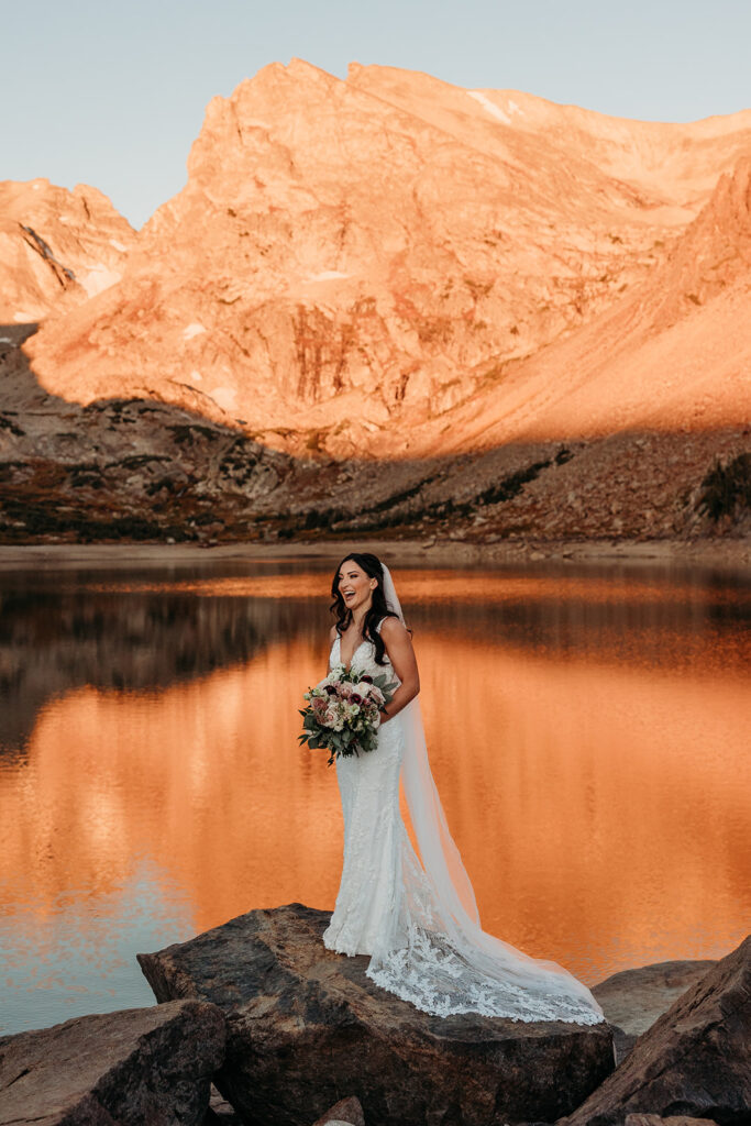 bride stands on the edge of a rock overlooking lake with sunrise