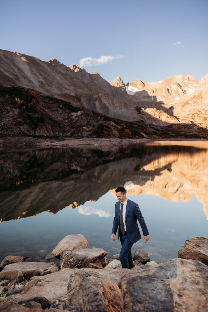groom walks up rocks on edge of a lake