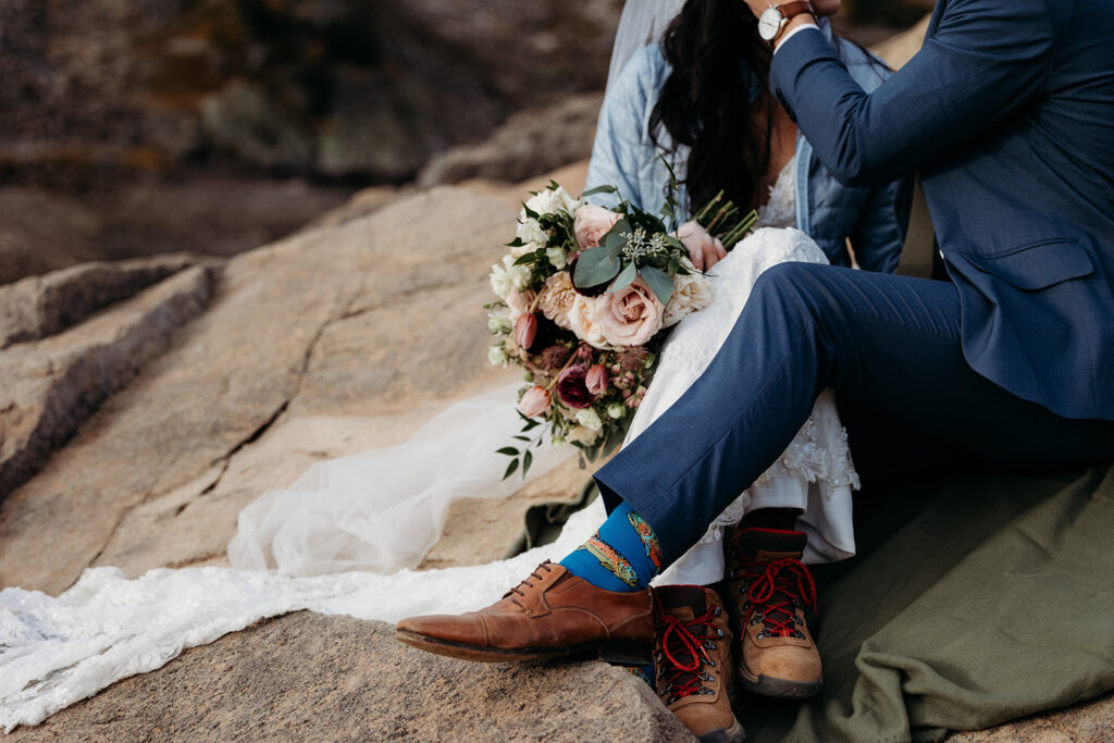 groom in blue suit with brown shoes sits on rock with bride