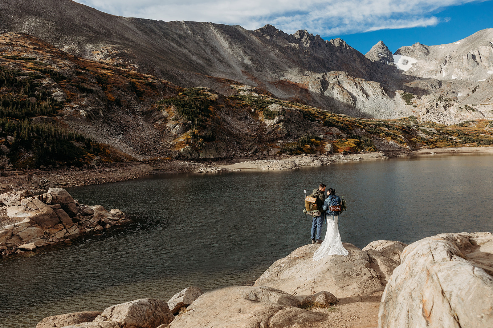 bride and groom wearing hiking backpacks in front of lake