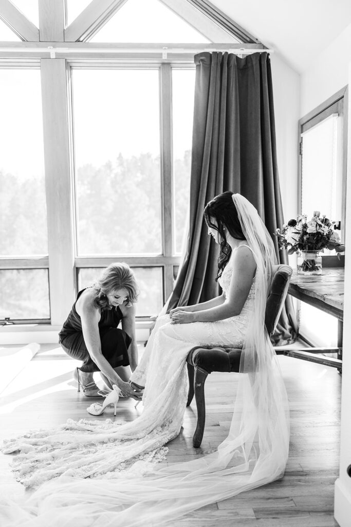 older woman helps put on brides shoes in front of windows