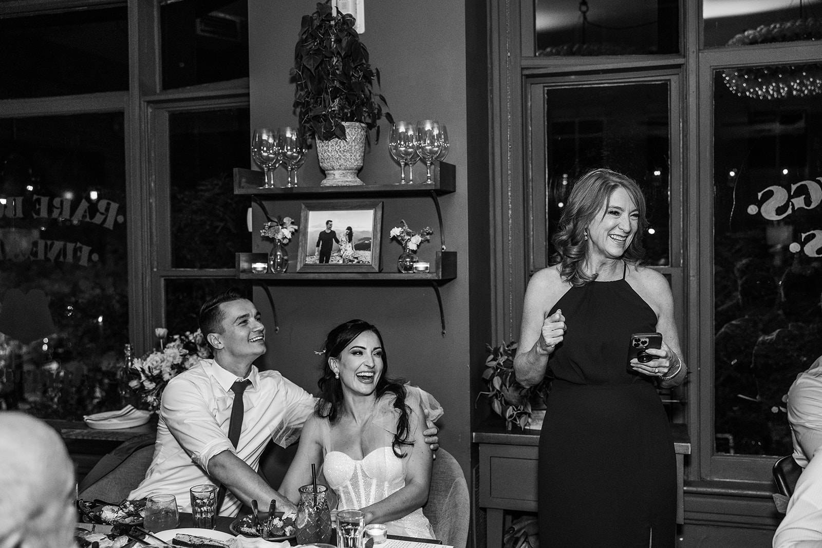 bride and groom look up at a woman who is standing holding a microphone next to their table